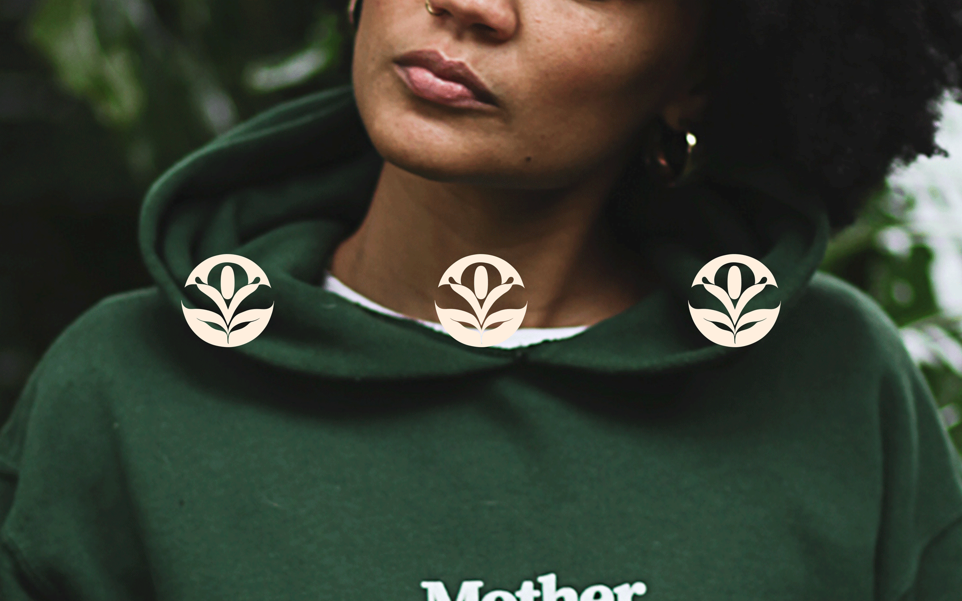 Mother of Plants Symbol in Light Deep Safron repeated 3 times in a row. Overlayed on photograph of Mother of Plants Founder in green hoodie. Designed by Can O' Baked Beans 2020